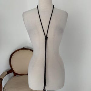May include: A black beaded necklace displayed on a mannequin. The necklace has a long, slender design with a knot detail at the center and a tassel at the end. The mannequin is set against a white background.