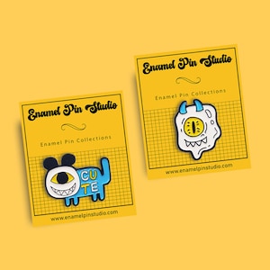 May include: Two enamel pins on a yellow background. The first pin is blue and white with a cartoon monster with a big eye and the text "CU TE". The second pin is white with a cartoon monster with a big eye and a mouth with teeth. Both pins have the text "Enamel Pin Studio" and "Enamel Pin Collections" on the background.