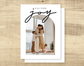 Puede incluir: Una tarjeta de felicitación blanca con las palabras "IN ALL THINGS joy" en escritura negra. La tarjeta presenta a una familia abrazándose dentro de un arco. La familia incluye dos adultos y dos niños. El fondo es una ventana con vistas.