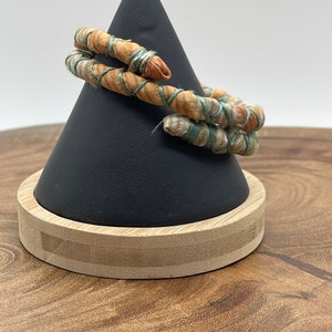 May include: A spiral bracelet with a unique design. The bracelet is made of a light brown material wrapped with a teal-colored thread. The bracelet is displayed on a black cone-shaped stand with a wooden base.