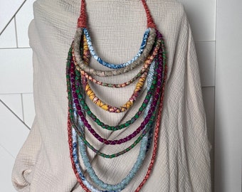 Puede incluir: Un collar de múltiples hilos con una variedad de hilos envueltos en tela de colores. El collar presenta hilos en tonos de rojo, azul, verde, amarillo y morado. El collar se exhibe sobre una cabeza de maniquí negra sobre un fondo neutro.
