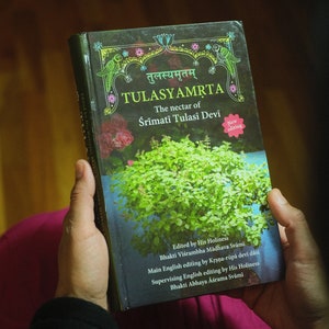 May include: A book titled "TULASYAMRTA" with the subtitle "The nectar of Śrīmati Tulasi Devi" in a dark green cover with a picture of a green plant and a pink flower. The book has a pink sticker that says "New editing".