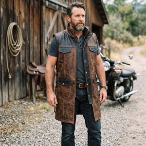 May include: A long brown leather vest featuring black leather accents on the collar, shoulders, and pocket flaps. The vest includes a front button closure, side lacing details, and four exterior pockets, worn over a dark gray shirt and blue jeans.