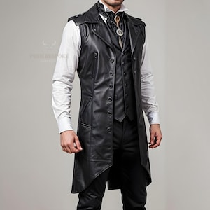Op de afbeelding: Een lange, zwarte leren vest met een getailleerde pasvorm. Het heeft een knoopsluiting, revers en zakken. Het vest wordt gedragen over een wit overhemd en een zwarte broek. De stijl is een mix van klassiek en modern.