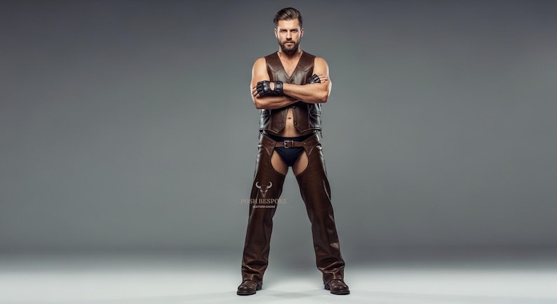 May include: A brown leather vest and chaps outfit. The vest has a V-neck and the chaps are open in the front. The outfit is worn with black gloves and boots. The model is standing with arms crossed.