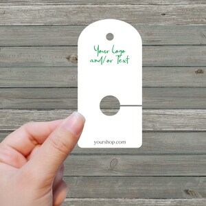 May include: White blank garment tag with a hole for hanging. The tag has a rounded top and a straight bottom. The text "Your Logo and/or Text" is written in green on the tag. The text "yourshop.com" is printed at the bottom of the tag.