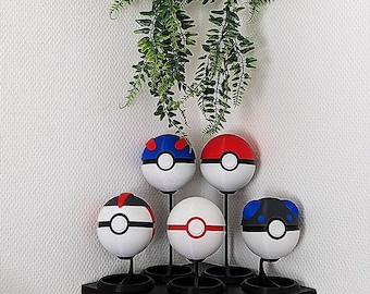 Op de afbeelding: Vijf Poké Balls op een zwarte standaard met een groene varen die erboven hangt. De Poké Balls zijn rood, blauw, zwart, wit en rood-wit.