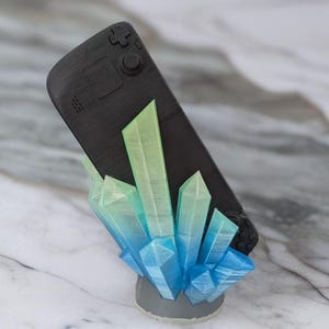 Peut inclure: Une console de jeu portable noire avec un support en forme de cristal vert et bleu. Le support est en plastique imprimé en 3D et présente un dégradé de couleurs du vert au bleu.