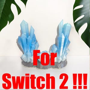 Peut inclure: Deux formations de cristaux bleus avec une base grise sont présentées sur un fond blanc. Le texte "For Switch 2 !!!" est superposé en rouge. Des feuilles vertes encadrent le haut de l'image.