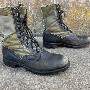 May include: Pair of well-worn combat boots. The boots feature a combination of olive green canvas and black leather. They have a lace-up design with metal eyelets and a sturdy, lugged sole. The boots show signs of wear and tear.