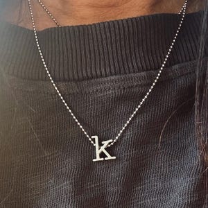 May include: A silver-colored necklace with a small letter "K" pendant. The pendant hangs from a delicate beaded chain. The necklace is displayed against a dark gray background.