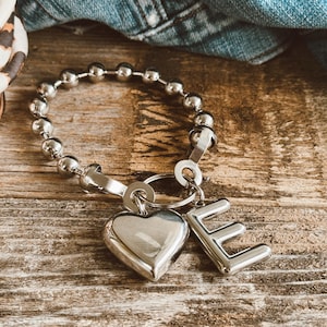 May include: A silver charm bracelet with a heart and the letter E charms. The bracelet is made of a chain of silver beads.