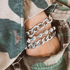 May include: Three silver chain bracelets stacked on a wrist. The top bracelet is a thick, chunky chain. The middle bracelet is a chain with large silver beads. The bottom bracelet is a thinner, more delicate chain.