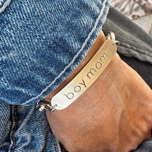 May include: A gold-colored bracelet with the words "boy mom" engraved on a rectangular plate. The bracelet is attached to a silver chain and is worn on a wrist. The background includes denim fabric.