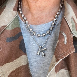 May include: A layered silver necklace with a ball chain and a pendant shaped like the letter "K". The necklace is worn over a gray t-shirt and a camouflage jacket.