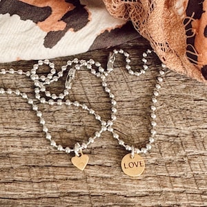 May include: Two silver ball chain necklaces with gold pendants. One pendant is a heart shape and the other is a round disc with the word "LOVE" engraved on it.