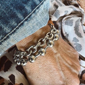 May include: A silver bracelet with two strands. One strand features a chain link design, and the other has a series of silver spheres. The bracelet is worn on a wrist, with denim and a leopard print fabric in the background.