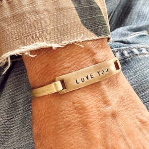 May include: A gold-toned metal cuff bracelet featuring a flat rectangular center plate with the words LOVE YOU stamped in black capital letters. The bracelet has a brushed finish and is connected to a thin band that wraps around the wrist.
