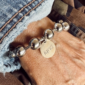 May include: Silver beaded bracelet with a round charm that says "kind". The bracelet features large, shiny, spherical beads strung together. The charm is a light gold color. The bracelet is worn on a wrist.