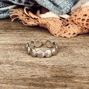 May include: A silver-toned metal ring featuring a continuous band of small, textured seashell shapes. The ring has an open-ended design, allowing for adjustable sizing, and displays a polished finish that reflects light across the shell details.