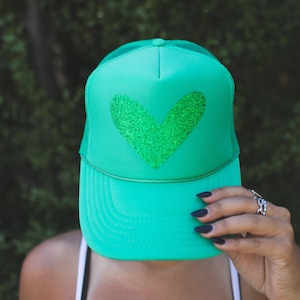 May include: A bright green trucker hat with a glittery green heart design on the front. The hat has a mesh back and a curved brim. The hat is a vibrant accessory.
