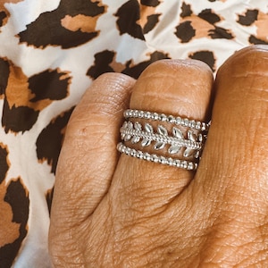 May include: A silver ring with a delicate leaf design and a beaded band. The ring is worn on a finger.