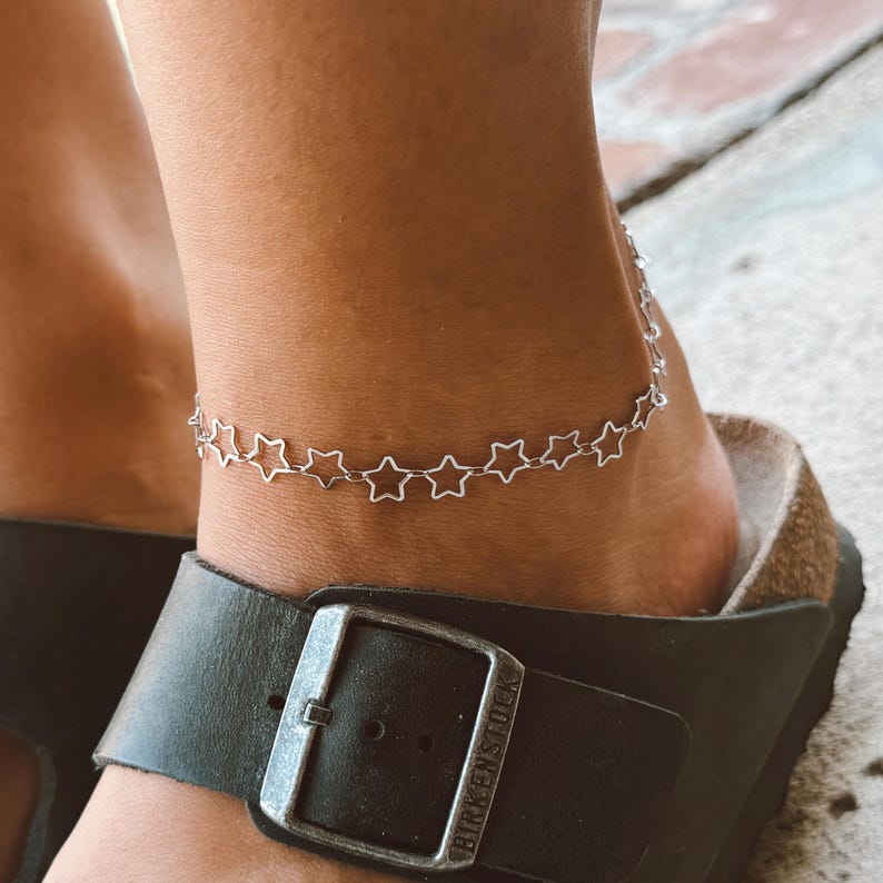 May include: A silver-colored star-shaped anklet is worn on an ankle. The anklet is made of small, connected star shapes. The ankle is above a black leather sandal with a silver buckle.