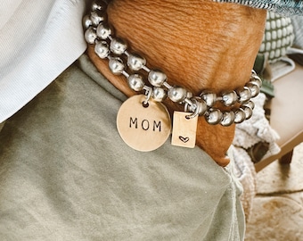 May include: A double-strand bracelet featuring metallic silver-toned beads. Two gold-toned metal charms are attached to the bracelet: a circular disc stamped with the text MOM and a smaller rectangular tag featuring a stamped heart symbol.