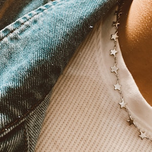 May include: A silver star chain necklace is draped over a white waffle-knit shirt. A blue denim jacket is visible in the upper left corner. The necklace is composed of small, linked star charms.