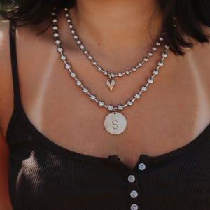 May include: Two-strand necklace with silver-colored beads. The top strand has a small heart pendant. The bottom strand features a circular pendant with the letter "S". The necklace is worn over a dark-colored tank top.