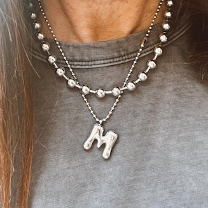 May include: A silver necklace with two strands. The top strand features small, round beads, while the bottom strand has a pendant in the shape of the letter "M". The necklace is worn over a gray t-shirt.