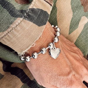 May include: A silver bracelet with a heart charm. The bracelet is made of round beads and is worn on a person's wrist.
