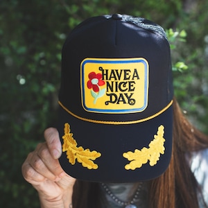 May include: Black trucker hat with a yellow patch that says "Have a Nice Day" with a red flower graphic. The hat has gold embroidered oak leaves on the brim.