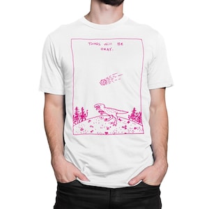 Puede incluir: Camiseta blanca con un gráfico rosa de un dinosaurio y un cometa que cae. El texto "Things will be okay." está por encima del dinosaurio.