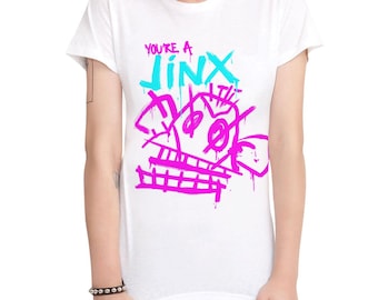 May include: White t-shirt with a pink and blue graphic design that says "You're a Jinx".
