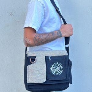 May include: Black and gray messenger bag with a white spiral sun design on the front pocket. The bag has a beige flap, a wooden ring detail, and a black adjustable strap. The bag is worn over the shoulder.