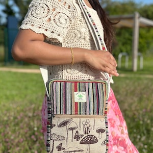 May include: A crossbody bag with a striped top section and a mushroom and butterfly print on the bottom. The bag has a hemp leaf logo and a cream-coloured strap. The person is wearing a cream-coloured crochet top.