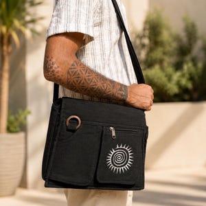 May include: A black canvas messenger bag with an adjustable shoulder strap, featuring two front pockets, a circular wooden ring attachment, and a white embroidered sun spiral graphic on the right pocket, worn across the body over a striped shirt.