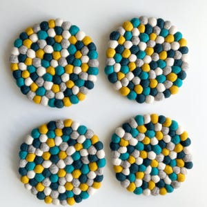 Puede incluir: Cuatro posavasos redondos de fieltro. Cada posavasos está hecho de pequeñas bolas de fieltro de colores en tonos turquesa, amarillo, gris y blanco. Los posavasos están dispuestos sobre una superficie blanca.