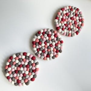 Può includere: Tre sottobicchieri rotondi in feltro nei toni del rosso, rosa, grigio e bianco. Ogni sottobicchiere è composto da piccole palline di feltro fitte. I sottobicchieri sono disposti su una superficie bianca.