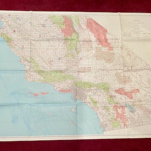 May include: A folded vintage map of California, featuring detailed geographical information. The map displays a light blue ocean, green and red shaded areas, and a network of roads and cities. The map is on a white background.