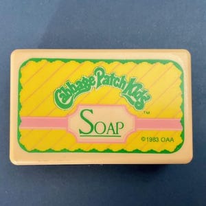 May include: A rectangular bar of soap with a light beige border. The soap features a yellow and green design with the words "Cabbage Patch Kids" and "SOAP" in a pink and green font. The copyright date is 1983.