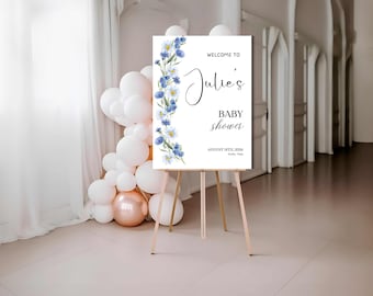 Op de afbeelding: Een wit bord op een houten ezel, met de tekst "Welcome to Julie's Baby Shower" en bloemmotieven. Het bord is versierd met blauwe bloemen en groen. Een tros witte en roségouden ballonnen staat op de achtergrond.