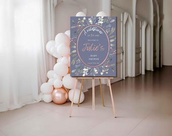 Op de afbeelding: Een babyborrelbord op een houten ezel. Het bord is stoffig blauw met bloemaccenten en de tekst "A wildflower in the making, Welcome to Julie's Baby Shower, August 19th, 2024" in roségoud.