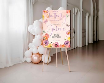Op de afbeelding: Een welkomstbord voor een babyshower op een houten ezel. Het bord is lichtroze met bloemen- en vlinderillustraties. De tekst luidt "A little butterfly is on the way! Welcome to Julie's Baby Shower August 18th, 2024."
