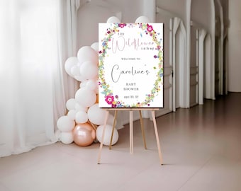 Op de afbeelding: Een welkomstbord voor een babyshower op een houten ezel. Het bord heeft een bloemenkrans met de tekst "A little Wildflower is on the way!" en "Welcome to Caroline's BABY SHOWER August 10th, 2021". Witte en roségouden ballonnen staan op de achtergrond.