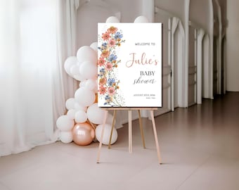 Op de afbeelding: Een wit bord op een houten ezel met de tekst "Welcome to Julie's Baby Shower" en bloemmotieven. Het bord is versierd met roze, blauwe en gele bloemen. Links staat een tros witte en roségouden ballonnen.