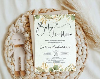 May include: A baby shower invitation with the text "Baby in bloom" in elegant script, adorned with botanical illustrations. The invitation includes details for Julie Anderson's baby shower, with the date, time, and location. A plush bunny toy with a tan scarf rests beside the invitation.