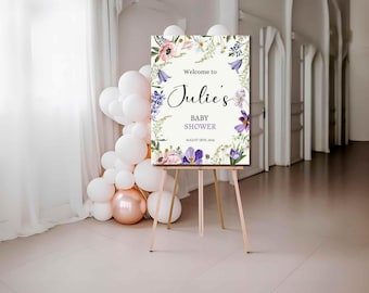 Op de afbeelding: Een houten ezel toont een welkomstbord voor een babyshower. Het bord toont de tekst "Welcome to Julie's BABY SHOWER" omringd door bloemenillustraties in roze, paars en wit. Links staat een cluster van witte en roségouden ballonnen.