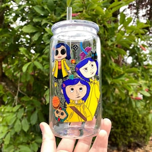 May include: A clear glass tumbler with a lid and straw, decorated with a colourful cartoon graphic. The design features three characters in yellow raincoats and blue hair. The background is a lush green with foliage.